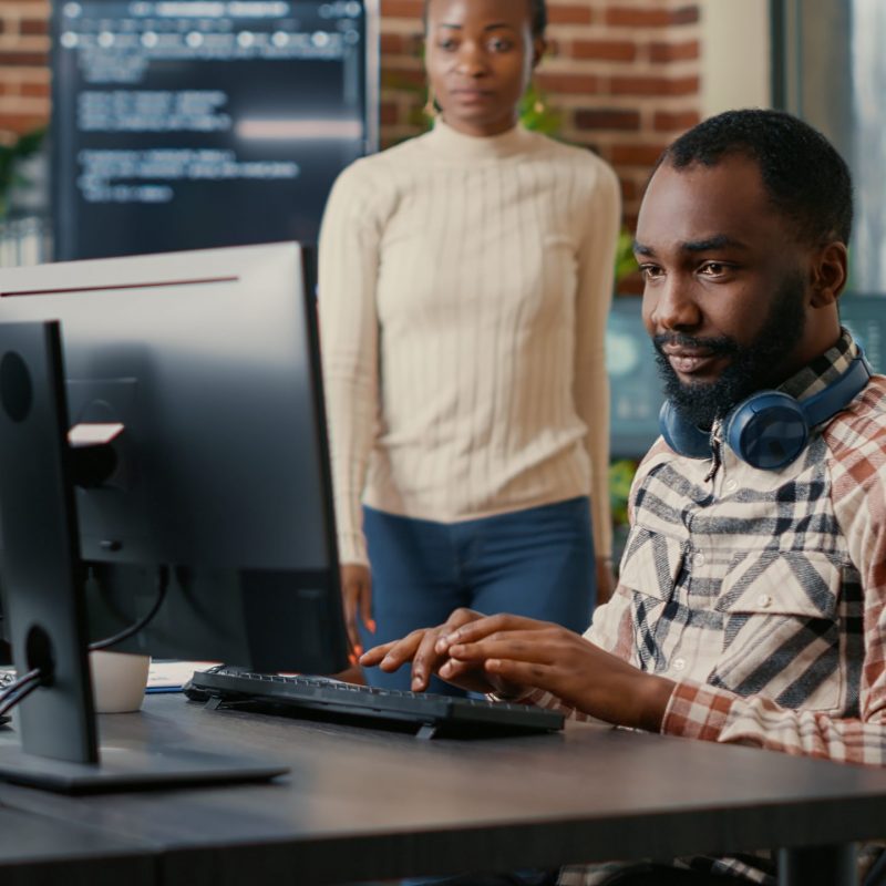 Relaxed software developer wearing wireless headphones smiling working looking at computer screen while typing. Coder writing code in it agency office with colleague asking for informations.