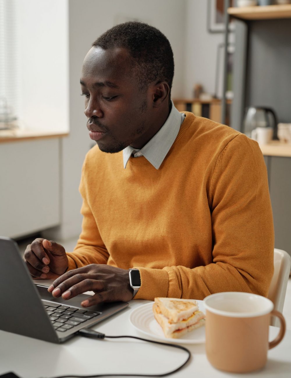 Programmer having lunch at his workplace African American programmer working on laptop and eating sandwich with coffee at his workplace at office