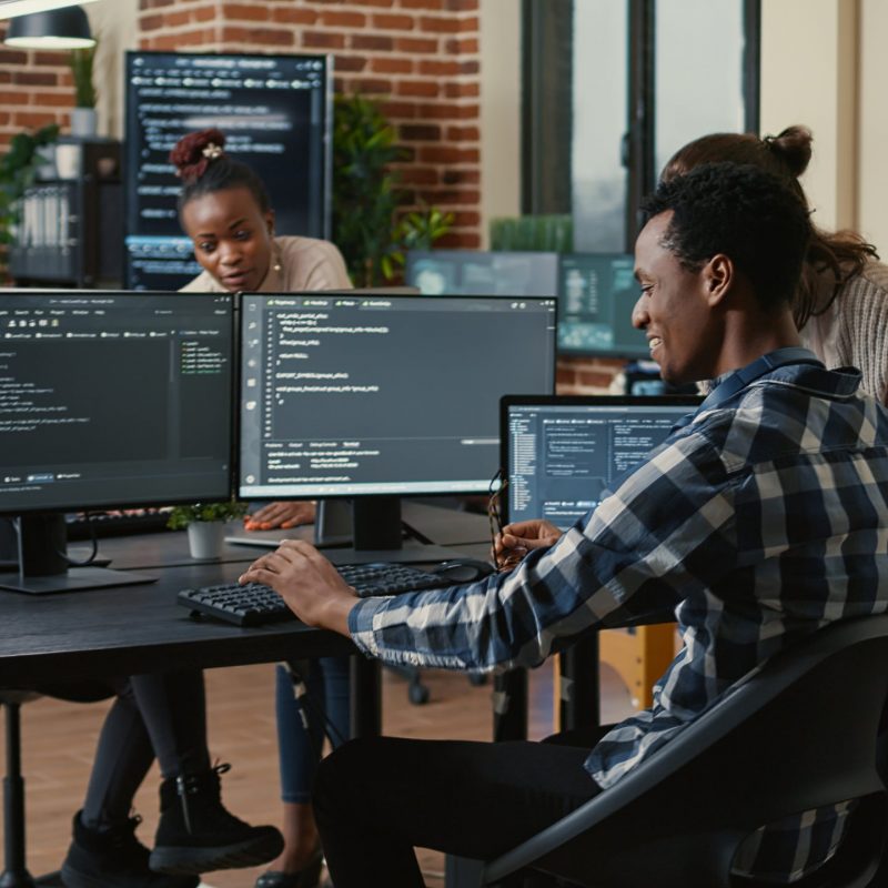 Software developer looking at running code on multiple screens takes off glasses and doing high five hand gesture with colleague programer. Programmers celebrating successful online cloud computing.